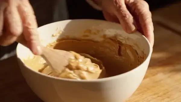 A wooden spoon folding thick pale almond paste over on itself in a white bowl