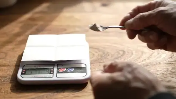 A hand tapping a very small heap of pale powder onto folded paper on a kitchen scale
