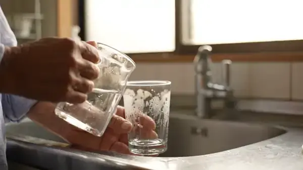 A hand pouring water from a jug into a glass at a kitchen sink in morning light